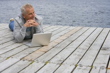 Senior man lying on pier in front of laptop, smiling