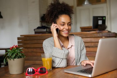 Smiling young woman using laptop and talking on mobile phone