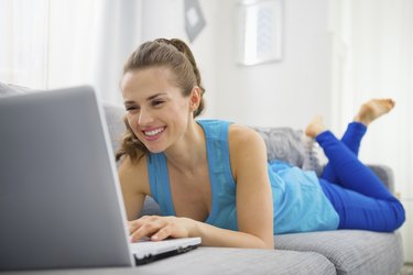 happy young woman laying on divan and using laptop