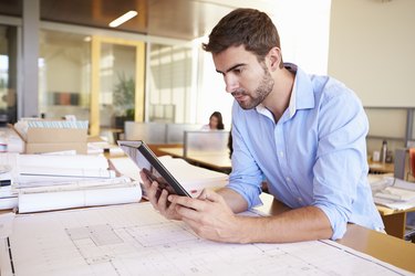 Male Architect With Digital Tablet Studying Plans In Office