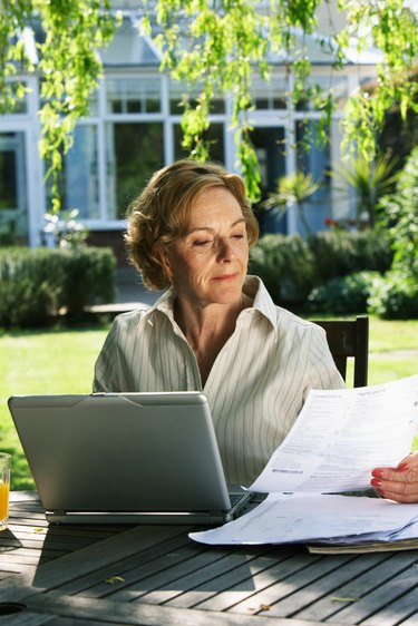 Senior woman sitting in garden with laptop, looking at paperwork