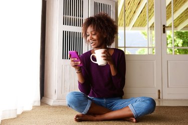 Smiling woman sitting on floor at home with cell phone