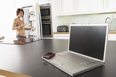 Young Woman Fixing Snack In Kitchen With Laptop In Modern Kitchen