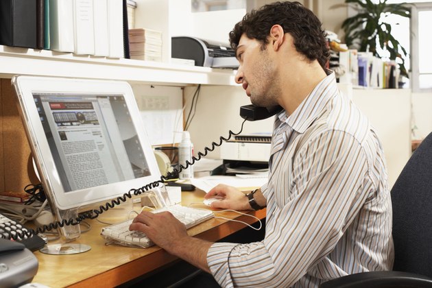 Male office worker balancing phone on shoulder, looking at computer