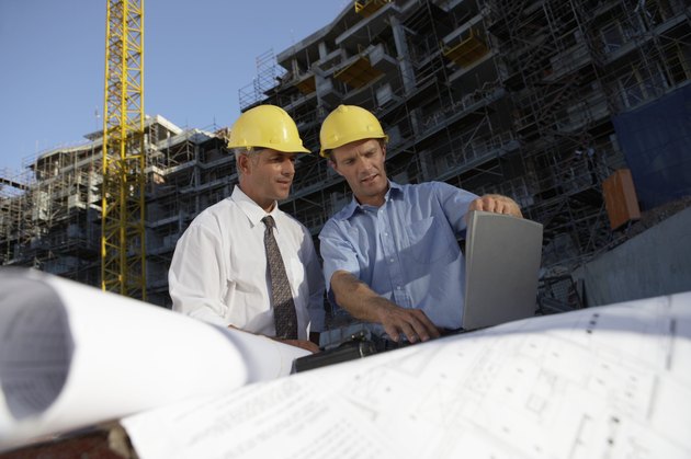 Two Men Wearing Hard Hats Looking Down at a Laptop Computer on a Building Site